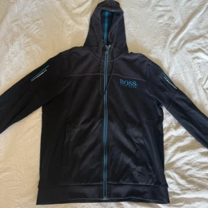 Svart zip kofta från HUGO BOSS storlek XL/L - Svart zip kofta från Hugo Boss i storlek XL men passar verkligen L också med blå logga och blå dragkedja. Tröjan har huva med snörning, långa ärmar och ribbade muddar. Tillverkad i mjukt material, perfekt för chill dagar. Regular fit och snygga blå detaljer på ärmarna. Jätte litet slitage nere vid handleden som man inte alls tänker på.