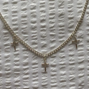 Silverfärgat halsband med korshängen - Stilrent halsband i silverfärg med tre små korshängen. Kedjan är av klassisk modell, lite tjockare än en vanlig och hängena har en enkel men snygg design. Halsbandet är helt nytt och har aldrig använts.