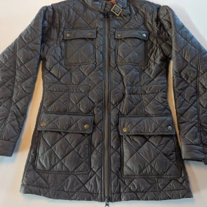 Quiltad jacka från Chevalier, brungrå - Snygg quiltad jacka från Chevalier i brungrå med orange foder. Jackan har fyra fickor framtill med tryckknappar, hög krage med spänne och dragkedja hela vägen. Perfekt för dig som gillar stilren och sportig look.