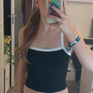 Svart och vit croppad linnetopp XS - Snygg croppad linnetopp i svart med vita kanter och smala axelband. Toppen har en enkel och stilren design som passar perfekt till jeans eller kjol. Tillverkad i mjuk och stretchig polyesterblandning för en bekväm passform.💞