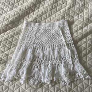 Vit virkad kjol med fransar - vit virkad kjol med fransdetaljer och knytband i sidan. Kjolen har ett bohemiskt och luftigt mönster. Fick den av en bekant och har själv aldrig använt den. Kan tänka mig att den är jättefin på stranden över en bikini, etikett finns inte vilket gör att jag inte vet storlek. Passar på mig som har storlek xs eller 158/164 på byxor och kjolar🥰