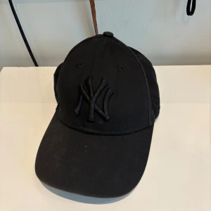 Svart New Era NY keps 9FORTY - Svart keps från New Era med klassisk New York Yankees-logga framtill i svart. Modellen är 9FORTY med böjd skärm och justerbar rem baktill. Tillverkad i bomull, perfekt för en sportig och stilren look.