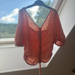 Blommig omlottopp i orange och vitt - Supersöt blommig omlottopp i orange och vitt med trekvartsärm och v-ringning. Toppen har snörning framtill och är gjord i ett lätt, luftigt material som passar perfekt till varma dagar. Den har en croppad passform och fina volangdetaljer längs nederkanten.