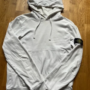 Vit hoodie från Stone Island - Säljer en clean vit hoodie från Stone Island med klassisk logga på ärmen. Tröjan har huva med snörning och en stor magficka. Tillverkad i mjuk bomull, perfekt för dig som gillar streetwear och vill ha en stilren look. Riktigt fet! STORLEK - sitter som S/M.