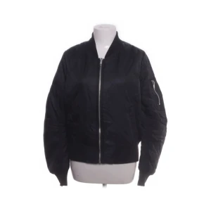 Svart bomberjacka med dragkedja - Stilren svart bomberjacka med klassisk siluett och silverfärgad dragkedja framtill. Jackan har ribbade muddar vid ärmslut och midja samt en ficka med dragkedja på ena ärmen. Perfekt för dig som gillar en clean och urban look.