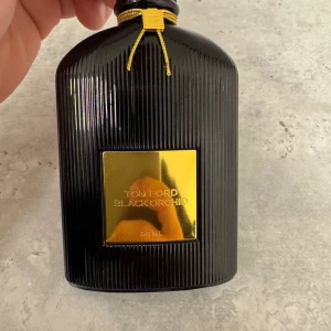 Tom Ford Black Orchid 50 ml parfym - Tom Ford Black Orchid Eau de Parfum i en lyxig svart, räfflad glasflaska med gulddetaljer och guldplakett framtill. Flaskan rymmer 50 ml och har en elegant, exklusiv design med guldfärgad snodd och TF-logga vid halsen.