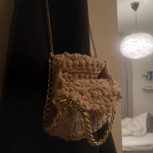 Beige/rosa stickad axelväska med kedja - Supersöt beige axelväska i stickat material med chunky guld-kedja som axelrem och detalj. Väskan har en mjuk, boxig form och är perfekt för att lyfta din outfit med en unik touch.