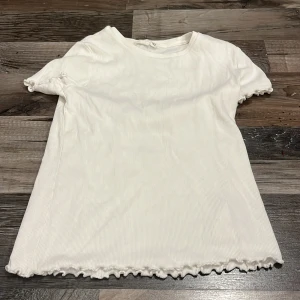 Vit ribbad topp från Kappahl - Söt vit ribbad topp från Kappahl med korta ärmar och volangkant vid ärmslut och nederkant. Enkel och stilren design som passar perfekt till jeans eller kjol. Mjuk och skön bomullskänsla, perfekt för en fräsch vardagslook.