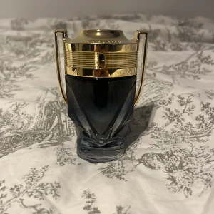 Paco Rabanne Invictus parfym - Snygg parfymflaska från Paco Rabanne Invictus, formad som en pokal med guldiga detaljer och mörkgrått glas. Flaskan har en modern och lyxig design som verkligen sticker ut på hyllan. Perfekt för dig som gillar exklusiva dofter och coola flaskor. Full flaska 50ml kan få med boxen om du vill! Snabbare affär = bättre pris‼️ kan byta mot en inviktus!