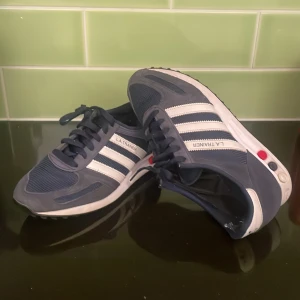 Adidas L.A. Trainer sneakers blå/vit - Snygga Adidas L.A. Trainer sneakers i marinblått och vitt med klassiska tre ränder på sidan. Ovandel i mocka och mesh, detaljer i vitt och färgade pluggar i sulan. Skorna har snörning och platt sula, perfekt för en sportig och retro look.