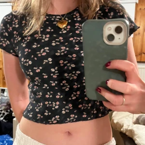 Blommig croppad topp från Pull&Bear - Supersöt croppad topp från Pull&Bear i marinblått med små blommor i vitt och rosa. T-shirten har korta ärmar och är ribbad i materialet, vilket ger en snygg och bekväm passform. Perfekt för dig som gillar en feminin och trendig stil! Har liten slitning under armen! Står storlek L men är mer en XS-S