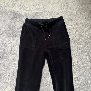 Svarta velourbyxor Juicy Couture Samt matchande Tröja - Svarta mjukisbyxor från Juicy Couture i mjuk velour med bootcut-ben och dragsko i midjan. Byxorna har stora fickor framtill och diskret broderad JC-logga på ena fickan. Perfekta för en chill och trendig look.