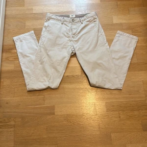 Beige slim fit chinos från Bowery - Snygga beige chinos från Bowery i slim fit-modell. Byxorna är tillverkade i mjuk bomull med lite stretch för extra komfort. Klassisk design med fickor fram och bak, perfekta för en clean och stilren look.