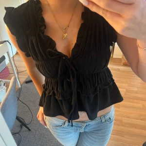 Svart blus med volanger från Gina Tricot - Supersöt svart blus från Gina Tricot med volangdetaljer och knytning framtill. Toppen har v-ringning, rynkade partier och är ärmlös med volangkant vid axlarna. Perfekt till jeans eller kjol för en trendig look.