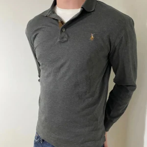 Mörkgrå pikétröja från Ralph Lauren - Snygg mörkgrå långärmad pikétröja från Ralph Lauren med klassisk krage och knappar framtill. Ikoniska broderade loggan på bröstet. Perfekt till jeans för en clean och stilren look.