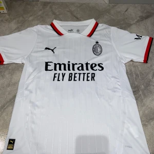 AC Milan bortatröja Puma Rafa Leão 10 - AC Milan bortatröja i vitt med svarta och röda detaljer, Puma-logga och klubbmärke på bröstet. Nummer 10 och Rafa Leão på ryggen. Kortärmad modell i lätt funktionsmaterial, perfekt för den varma sommaren.