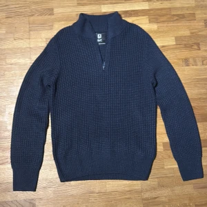 Mörkblå stickad tröja Brandit half zip - Mörkblå stickad tröja från Brandit med grov struktur och hög krage. Tröjan har en half zip-dragkedja framtill och långa ärmar. Perfekt för kyliga dagar och enkel att matcha med jeans eller cargobyxor.