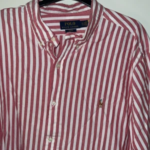 Polo Ralph Lauren skjorta  - Säljer en rosa och vit randig skjorta från Polo Ralph Lauren i slim fit. Skjortan har klassisk krage, knäppning framtill och broderad logga på bröstet. Tillverkad i bomull och har långa ärmar. Perfekt för dig som gillar stilren och tidlös design.