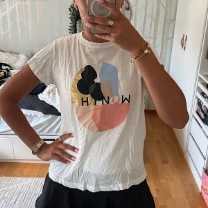 Vit t-shirt med grafiskt tryck från Munthe - Vit t-shirt från Munthe med coolt grafiskt tryck i guld, svart, blått och rosa på bröstet. Klassisk rund hals och korta ärmar. Skön passform och tillverkad i mjuk bomull. Perfekt för dig som gillar en stilren men ändå färgglad look. (Kan såklart strykas innan!)❤️