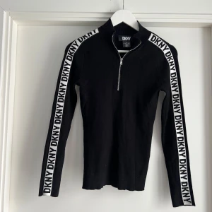 Svart ribbad DKNY tröja med dragkedja - Svart ribbad tröja från DKNY med vita logoband längs ärmarna och en snygg half zip-dragkedja framtill. Långärmad och tight passform, perfekt för dig som gillar sportig och trendig stil. Materialet är stretchigt och bekvämt.