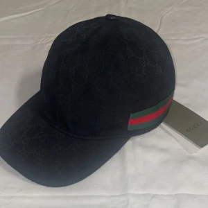 Gucci keps svart - Snygg svart keps från Gucci med diskret GG-monogram över hela tyget och klassiska gröna och röda ränder på sidan. Justerbar rem baktill i skinn och insida med märkning. Perfekt accessoar för en trendig look.