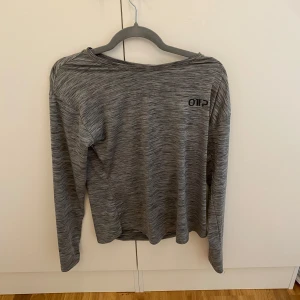 Grå långärmad träningströja OTP - Säljer en gråmelerad långärmad träningströja från OTP. Tröjan har en avslappnad passform och är tillverkad i ett lätt och syntetiskt material som andas. Perfekt för gymmet eller löprundan. Diskret logga på bröstet.