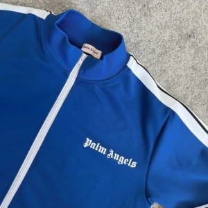 Blå tracksuit från Palm Angels - Blå track tröja från Palm Angels med vita ränder längs ärmarna och vit dragkedja. Tröjan har ståkrage, två fickor med dragkedja och logga på bröstet. Tillverkad i bomull och polyester för en sportig vibe.