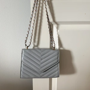 Ljusgrå quiltad axelväska med kedja - Snygg ljusgrå axelväska med quiltad design och guldfärgad kedjerem. Väskan har ett stilrent kuvertformat lock och är tillverkad i syntetmaterial. Perfekt för att lyfta din outfit med en trendig touch.