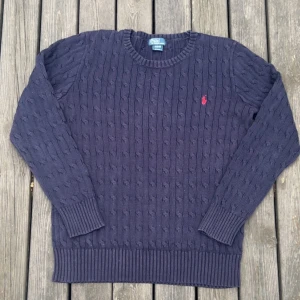 Mörkblå kabelstickad tröja Polo Ralph Lauren - Kabelstickad mörkblå tröja från Polo Ralph Lauren med klassisk rund hals och röd broderad logga på bröstet. Tröjan är i bomull och har ribbade muddar vid ärmslut och nederkant. Perfekt för dig som gillar stilren och tidlös look. Storlek XL för barn men passar perfekt på mig som är 172 cm lång.