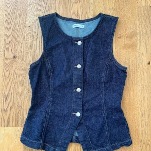 Mörkblå jeans top från Gina Tricot - Snygg mörkblå jeansväst från Gina Tricot med klassisk rund halsringning och silverfärgade knappar framtill. Västen har figurnära passform och markerade sömmar som ger en cool och tidlös look. Perfekt att styla över en t-shirt eller skjorta.