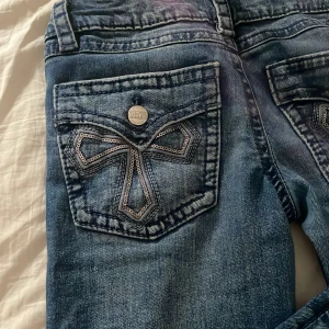 Blå jeans med paljettficka Gina Tricot - Snygga blå jeans från Gina Tricot i storlek 152. Jeansen har coola bakfickor med paljettbroderi i form av en rosett och tydliga kontrastsömmar. Klassisk femficksmodell med knapp och dragkedja. Perfekta för dig som vill sticka ut lite extra.