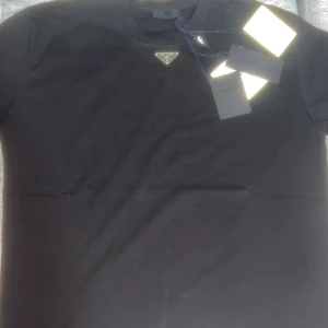 Svart t-shirt från Prada - Snygg svart t-shirt från Prada med klassisk passform och diskret logga på bröstet. T-shirten är enkel men exklusiv, perfekt för dig som gillar stilrena plagg med lyxig känsla. Tillverkad i mjuk bomull och har korta ärmar.