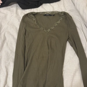 Olivgrön ribbad topp med spets Vero Moda - Olivgrön långärmad topp från Vero Moda med ribbad struktur och v-ringning dekorerad med blommig spets. Perfekt för dig som gillar stilrena detaljer och mjuka material. Snygg att matcha med jeans eller kjol för en chill look.