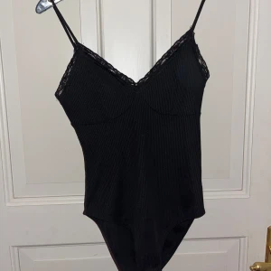 Svart body med spets - Snygg svart ribbad body med tunna axelband och v-ringning dekorerad med spets. Toppen har en figurnära passform och är perfekt för att skapa en stilren look. Materialet är mjukt och stretchigt för extra komfort.