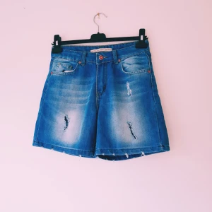 Blåa jeansshorts från Kilroy denim strl 28 - Snygga blåa jeansshorts från Kilroy med hög midja och slitna detaljer framtill och baktill. Shortsen har klassisk femficksdesign, knapp och dragkedja samt råa kanter nertill. Perfekta för en avslappnad och trendig look i sommar.  Helt & fint skick