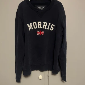 Mörkblå sweatshirt från Morris - Mörkblå sweatshirt från Morris med broderad logga och brittisk flagga på bröstet. Klassisk rund halsringning och långa ärmar. Perfekt för dig som gillar stilren och sportig look. Tillverkad i mjuk bomull för extra komfort.
