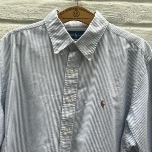 Randig skjorta från Ralph Lauren - Klassisk skjorta från Ralph Lauren i blåvitt randigt mönster. Skjortan har lång ärm, knappar framtill och broderad logga på bröstet. Classic fit-modell i mjuk bomull, perfekt för en clean och tidlös stil.