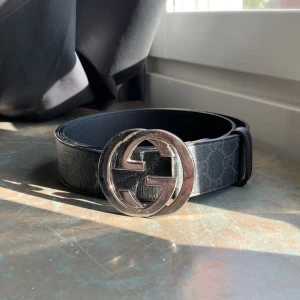 Svart Gucci bälte med GG-spänne - Snyggt svart bälte från Gucci med klassiskt GG-mönster och stort silverfärgat GG-spänne. Bältet är gjort i skinn och har en stilren, bred design som sticker ut. Perfekt för dig som vill ha en lyxig accessoar med ikonisk känsla.