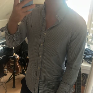Randig skjorta från Ralph Lauren - Säljer en klassisk skjorta från Ralph Lauren med smala blå och vita ränder. Skjortan har lång ärm, knappar hela vägen och den ikoniska broderade loggan på bröstet. Tillverkad i mjuk bomull och har en snygg, normal passform. Perfekt för dig som gillar stilrena plagg med preppy-vibe.
