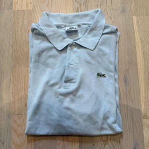 Ljusblå pikétröja från Lacoste - Snygg ljusblå pikétröja från Lacoste med klassisk krage och knäppning framtill. Ikoniska gröna krokodilloggan broderad på bröstet. Tillverkad i bomull för en skön och luftig känsla. Perfekt för dig som gillar stilren och sportig look.
