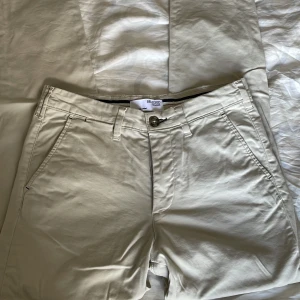 Beige chinos från Selected Homme - Snygga beige chinos från Selected Homme med klassisk passform. Byxorna har snedställda fickor fram, två bakfickor med knapp och bälteshällor. Tillverkade i mjuk bomull, perfekta för en clean och stilren look.
