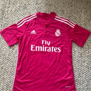 Real Madrid Ronaldo fotbollströja - Rosa Real Madrid fotbollströja från Adidas med Ronaldo och nummer 7 på ryggen. Tröjan har vita detaljer, klassiska Adidas-ränder på axlarna och Fly Emirates-tryck på bröstet. Tillverkad i lätt och ventilerande climacool-material. Perfekt för dig som älskar fotboll och Ronaldo. Storleken är S men kan passa mindre M