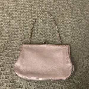 Silver kuvertväska med kedja - Stilren kuvertväska i metallic silver med kedjehandtag och klassiskt metallspänne. Väskan har en mjuk, glansig yta och ett ljust innerfoder med liten innerficka. 