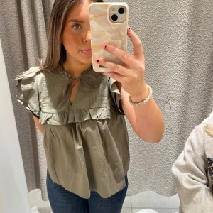 Olivgrön blus med volang och knyt - Säljer en olivgrön blus med volangdetaljer och knyt vid halsen. Blusen har korta ärmar med rynkade axlar och en luftig, loose passform. Perfekt för dig som gillar en söt och avslappnad stil.