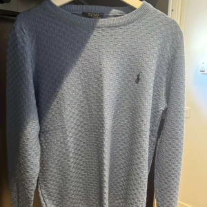 Ljusblå tröja från Polo Ralph Lauren - Säljer en ljusblå långärmad tröja från Polo Ralph Lauren med diskret rutmönster och broderad logga på bröstet. Tröjan har rund halsringning och är tillverkad i mjuk bomull, perfekt för en chill och stilren look.