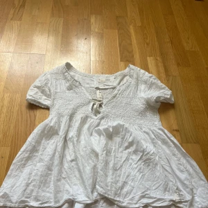 Vit blus med knyt från Odd Molly - Supersöt vit blus från Odd Molly med korta puffärmar, smock över bysten och v-ringning med knytband. Blusen har diskret prickigt strukturmönster i tyget och är tillverkad i mjuk bomull. Perfekt för en romantisk och avslappnad look.