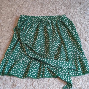 Grön blommig omlottkjol med volang - Supersöt grön kortkjol med vita blommor, omlottmodell och volangkant. Kjolen har knytband i midjan och är perfekt för sommarens alla äventyr. Lätt och luftig känsla, passar perfekt till sneakers eller sandaler.