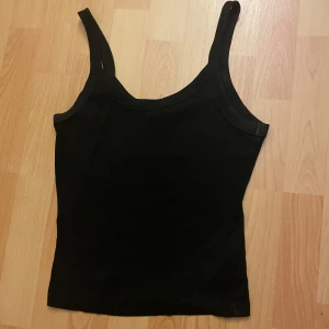 Svart ribbad linnetopp - Enkel och stilren svart linnetopp med breda axelband och rundad halsringning. Toppen är ribbad och sitter snyggt mot kroppen, perfekt att matcha med jeans eller kjol för en clean look.