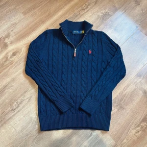 Marinblå kabelstickad tröja Polo Ralph Lauren - Kabelstickad marinblå tröja från Polo Ralph Lauren med halv dragkedja och hög krage. Tröjan har lång ärm, ribbade muddar och röd broderad logga på bröstet. Perfekt för dig som gillar klassisk och stilren look.