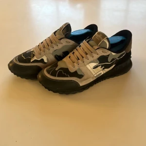 Valentino Rockrunner sneakers camo - Snygga Valentino Rockrunner sneakers med camouflage-mönster i svart, grått och beige. Skorna har svarta och beige partier, rund tå, snörning och en robust svart sula. Perfekta för dig som vill sticka ut med exklusiva detaljer och cool design.